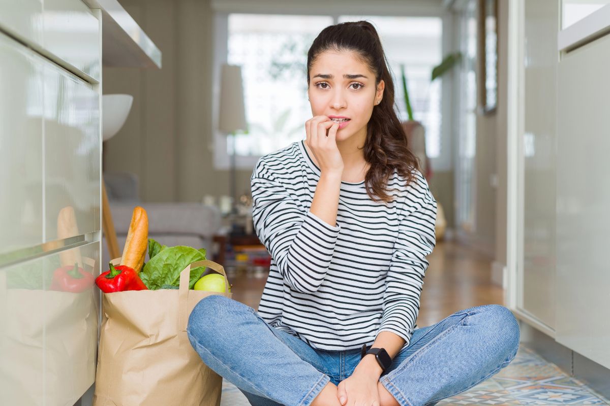 woman sitting on the kitchen floor looking worried with groceries in a bag near her