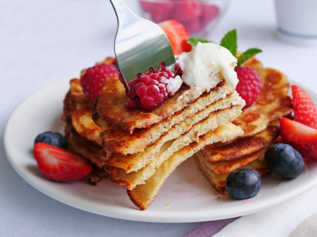 Keto pancakes made of coconut flour or almond flour, served with berries and whipped cream. Slice of stacked pancakes on fork.