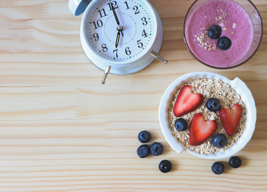 Top view or flat lay of alarm clock, breakfast with a glass of blueberry smoothie and oat or granola in white bowl, fresh blueberries, strawberries on wooden table. Healthy breakfast concept.