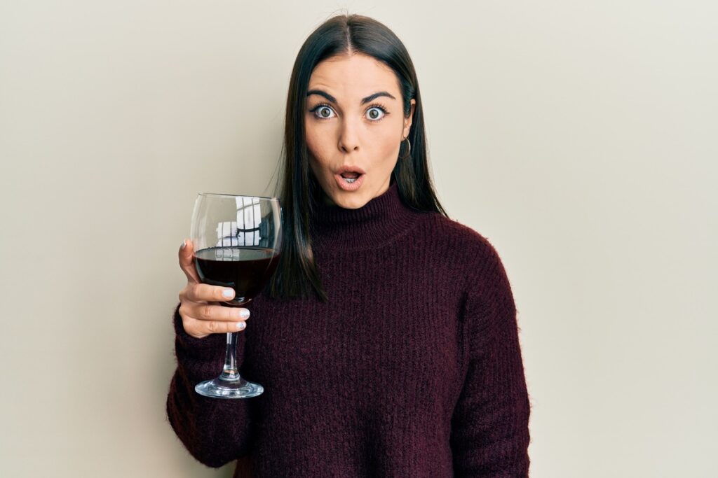 Young brunette woman drinking a glass of red wine scared and amazed with open mouth for surprise, disbelief face