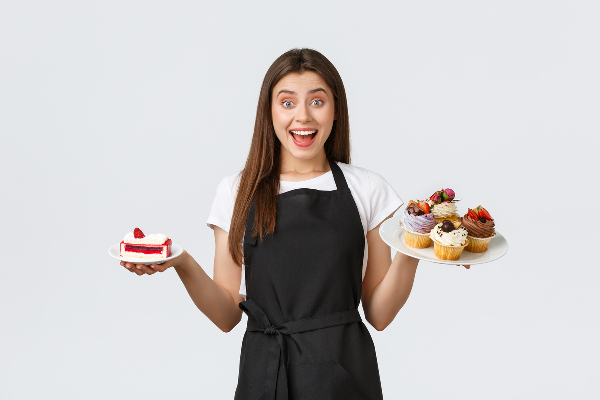 Grocery store employees, small business and coffee shops concept. Friendly smiling waitress in cafe holding plates with cupcakes and cafe, serving desserts for guests, standing white background.