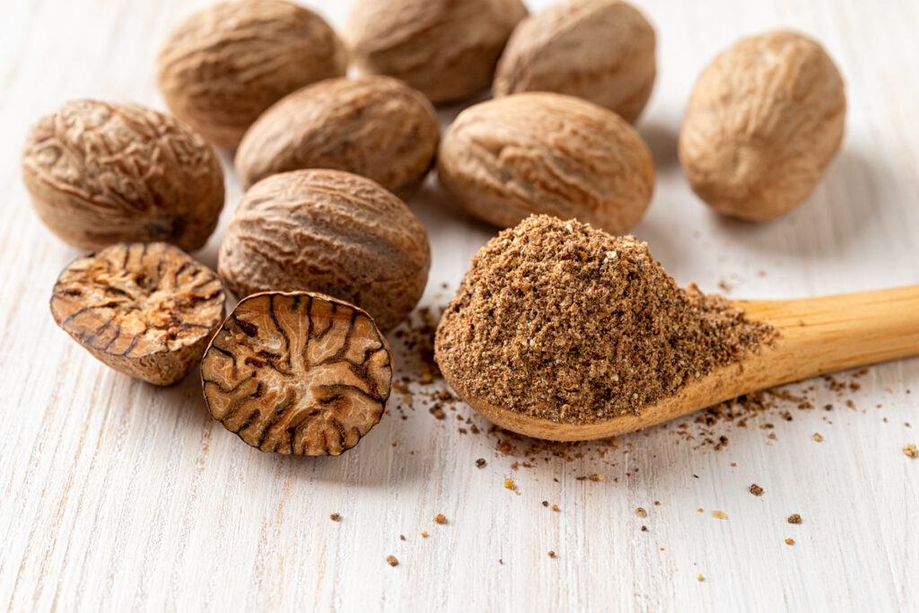 Spoon of ground nutmeg, whole and halved seeds over white wooden background. Muscat nuts closeup for spice and seasoning concept. Myristica fragrans tree organic fruits for herbal medicine. Front view.