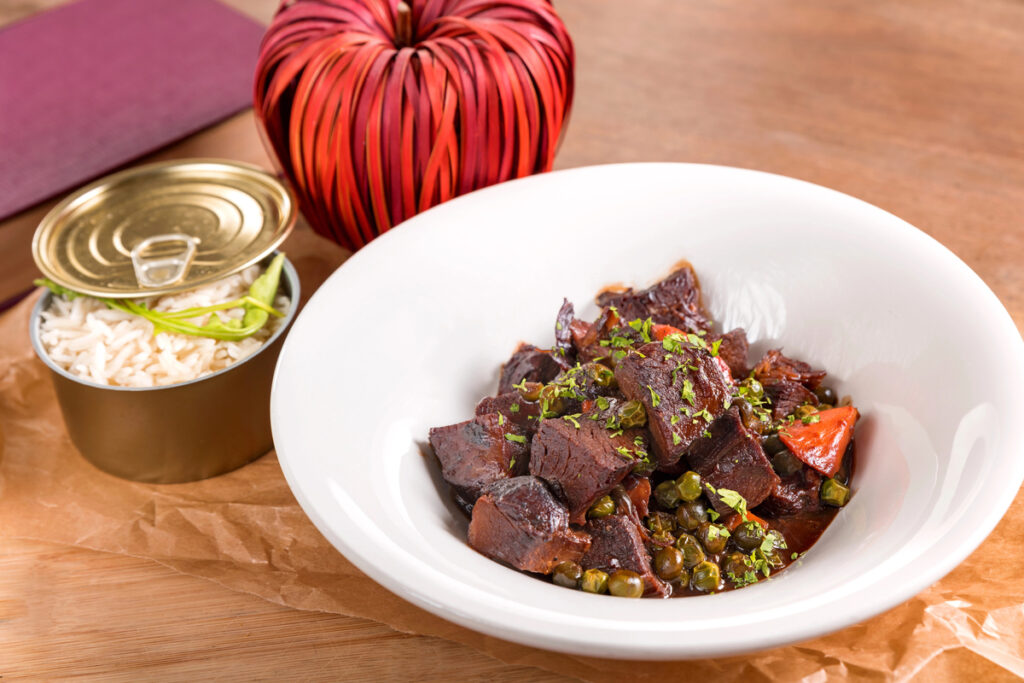 Stewed beef cheeks in red wine sauce with peas and carrots, garnished with white rice, served in a white plate on a wooden table..