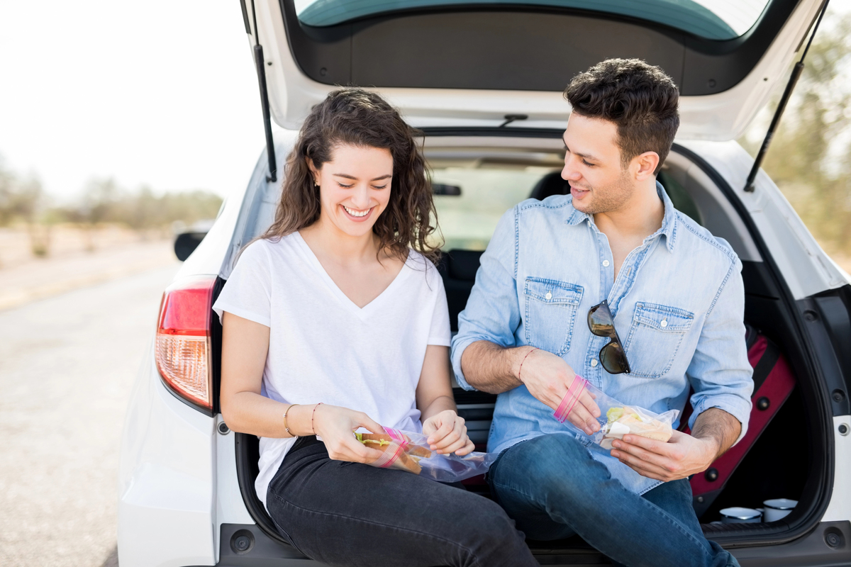 Happy young couple sitting together in a trunk of their car and eating sandwich during their road trip