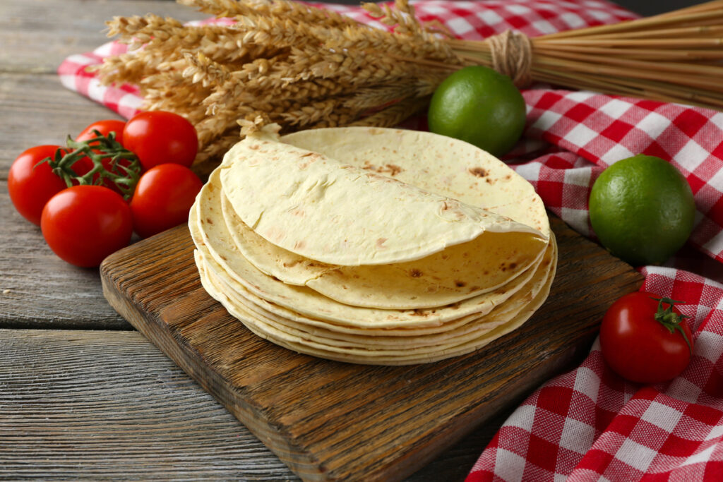 Stack of homemade whole wheat flour tortilla and vegetables on cutting board, on wooden table background