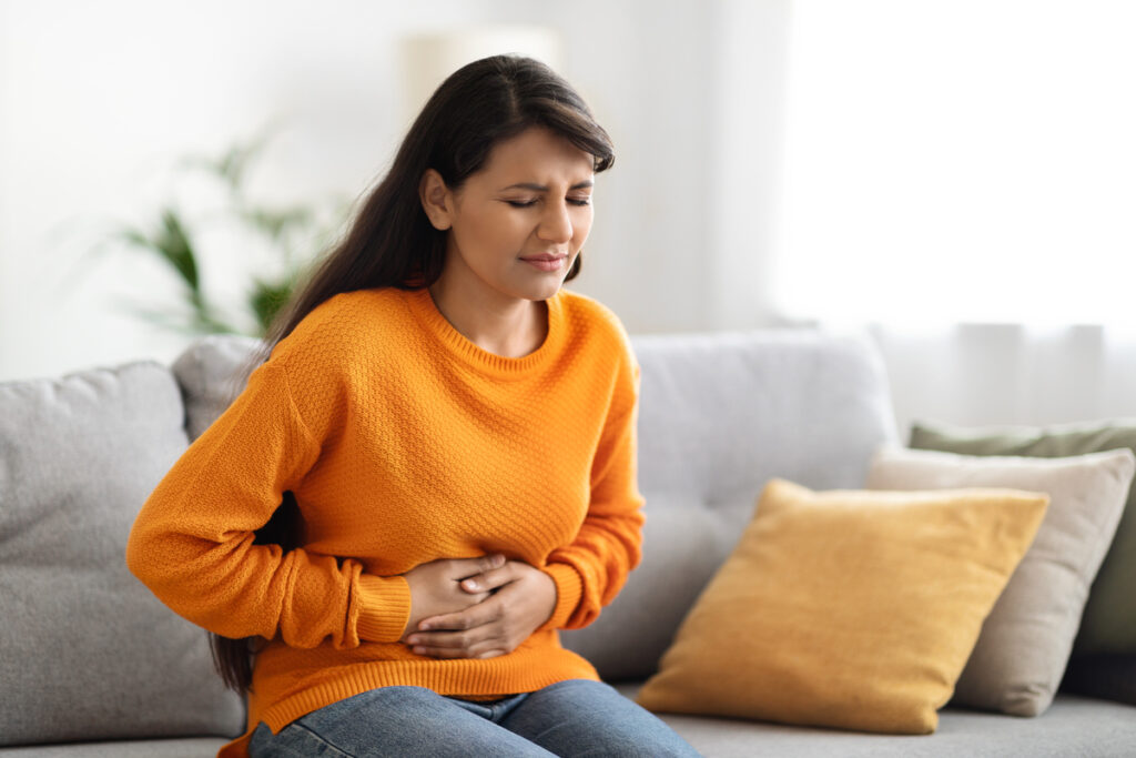 Concerned unhappy young indian woman in casual suffering from stomach-ache, sitting on sofa at home. Female holding tummy, undergoing belly pain and discomfort, suffer from menstruation, copy space