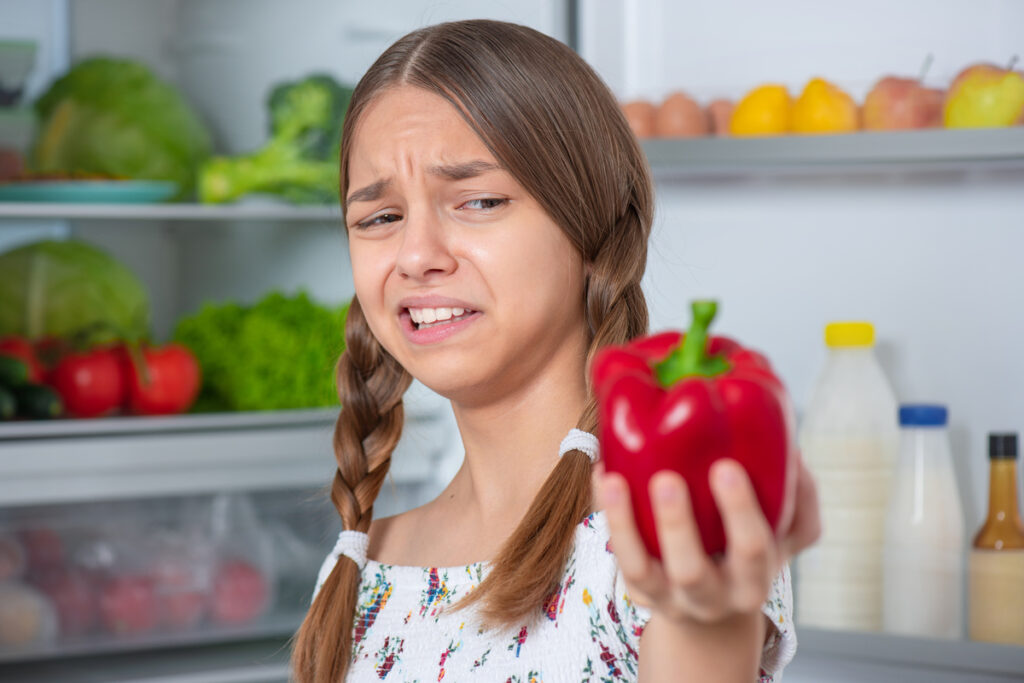 Teen girl holding fresh red pepper while standing near open fridge in kitchen at home. Child do not want to eat vegetables and dislike taste of pepper. Organic natural healthy food produce.