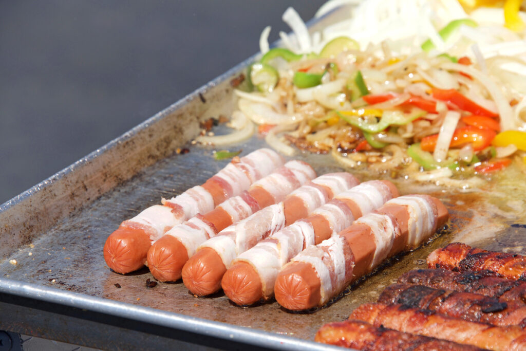 Street vendor cooking bacon wrapped hot dogs with onions and peppers grilling beside them. Popular cuisine for street fairs and events