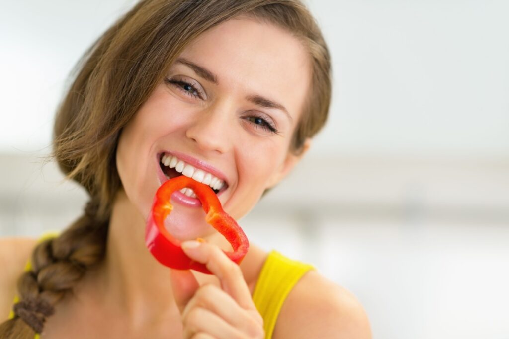 happy young woman eating bell pepper in kitchen