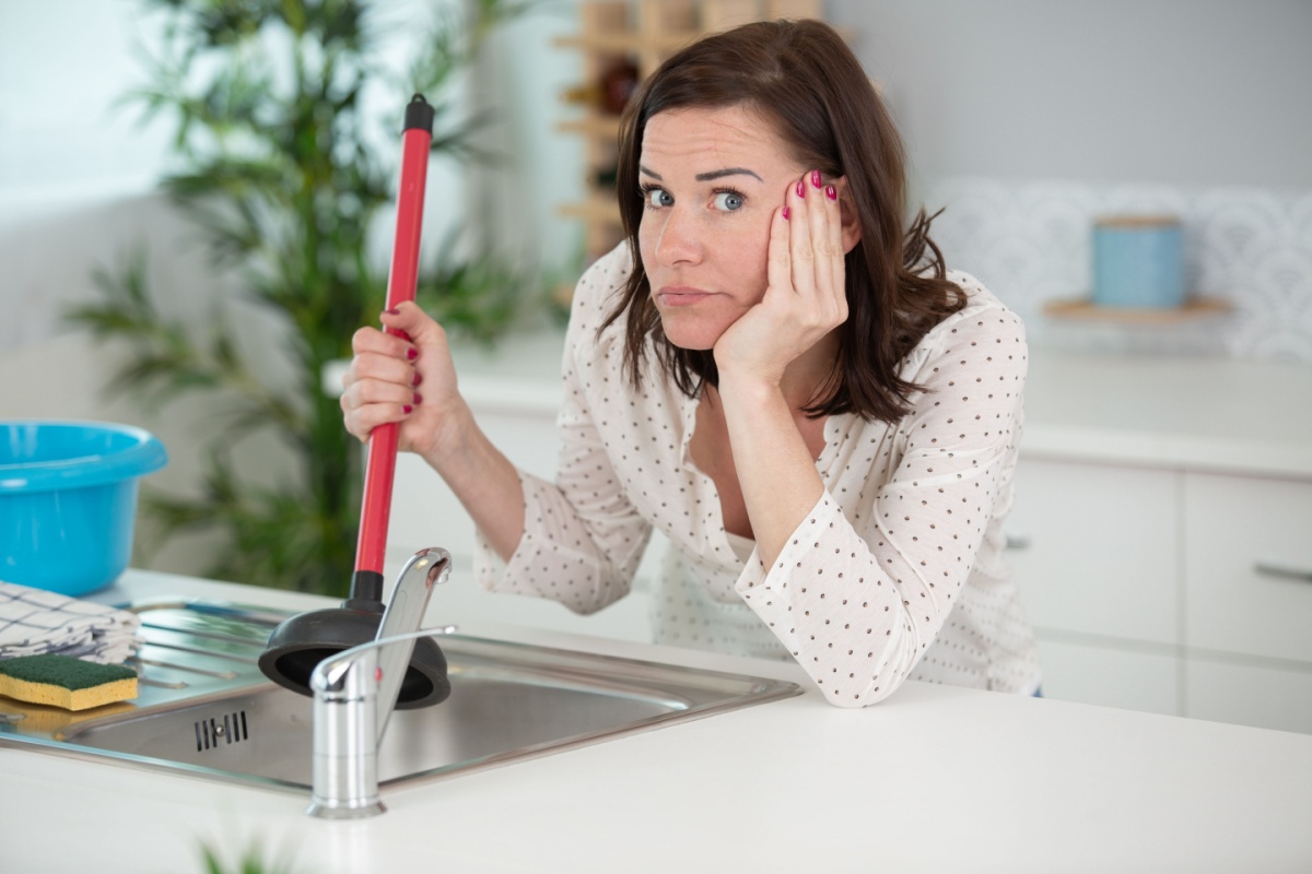 upset woman using plunger in blocked kitchen sink