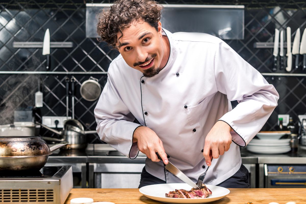 A chef in a kitchen smiling and cutting meat