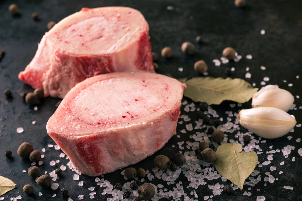 Horizontal photo with two pieces or slices of big marrowbone with few drops of blood and meat on side. Bones are placed on worn black baking tray with salt and other spices around.
