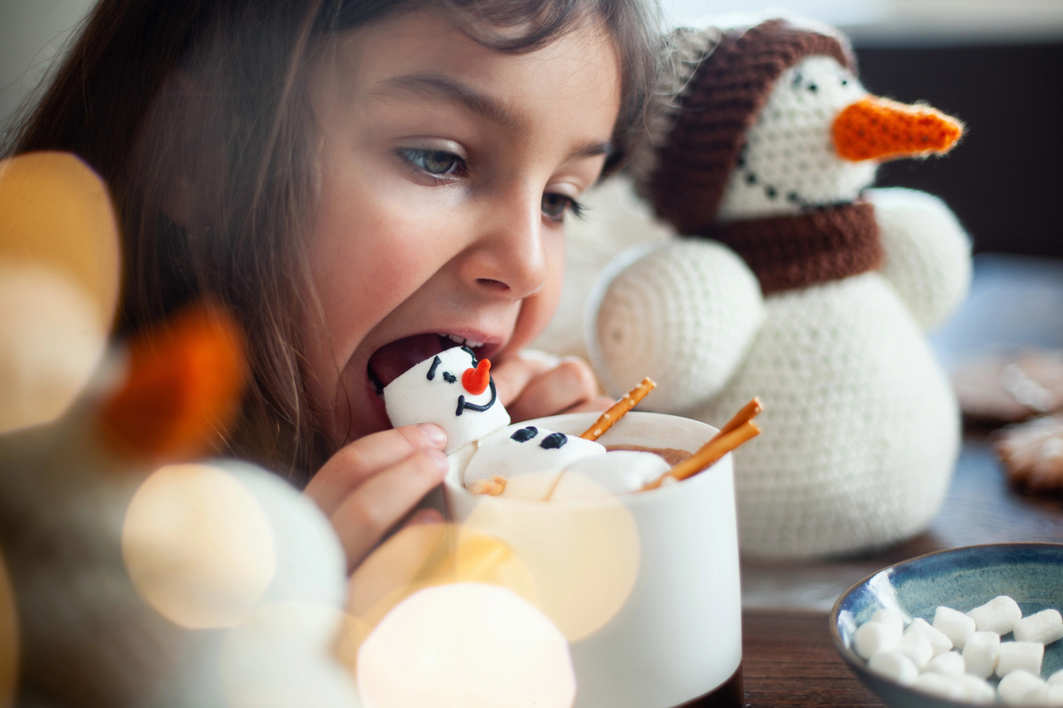 Little cute girl eats a marmmello snowman decorated with icing and drinks cocoa. Concept Preparing for Christmas and New Year.