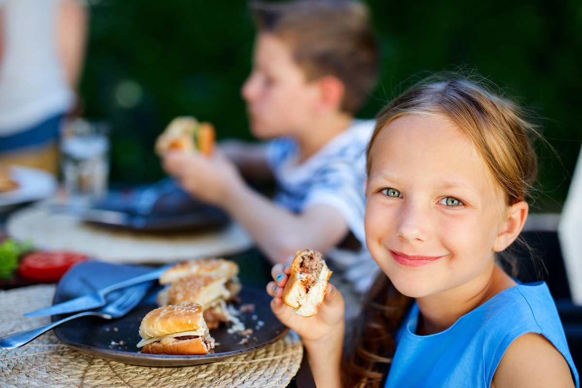 Adorable little girl and her family eating delicious homemade burger outdoors on summer day